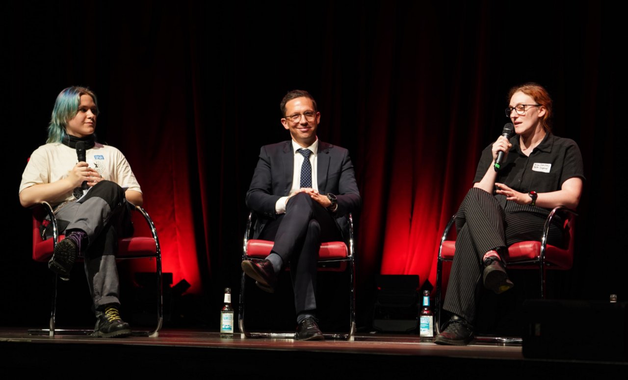 Das Bild zeigt die Frontalansicht einer bestuhlten Bühne, auf der drei Personen von links nach rechts abgebildet sind: Die Freiwillige Luna Machens, die das Podiumsgespräch moderiert, Kulturminister Falko Mohrs, der direkt in die Kamera blickt und lächelt, und Kimberby Haarstik vom Deutschen Gewerksschaftsbund Jugend, die gerade ins Mikrofon spricht. 