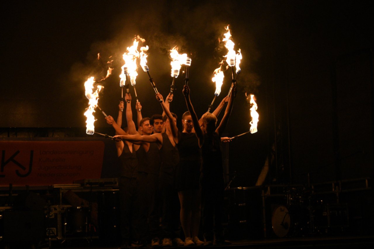 Das Bild ist in der Dunkelheit aufgeommen worden. Auf der Bühne stehen hintereinander aufgereiht junge Frauen und Männer, ganz in schwarz gekleidet, die brennende Fackeln in alle Richtungen halten.