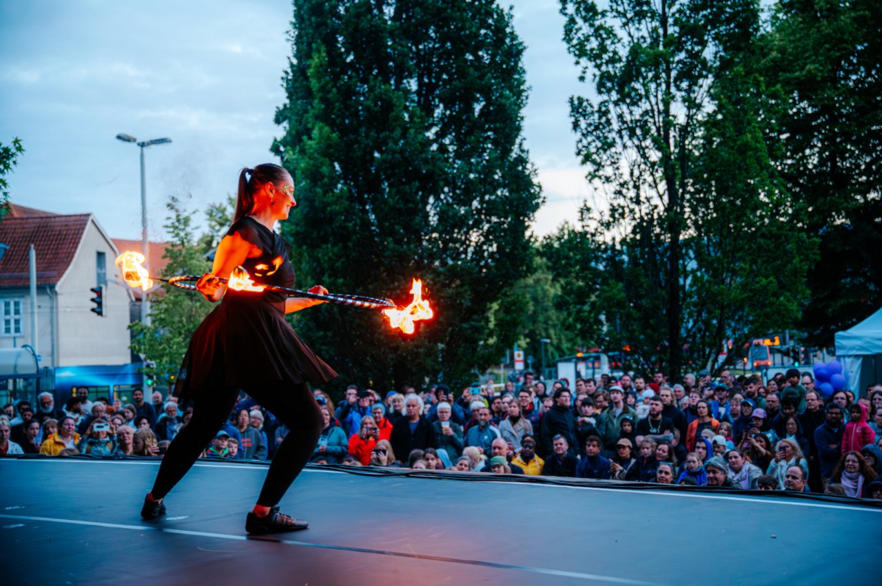Das Bild zeigt von der hinteren Bühne aus einen Blick ins Pubikum, das in der Abenddämmerung sitzt. Auf der Bühne steht Lana Ebel von Artistikka aus Damme und schwingt gerade einen brennenden Hullahop-Reifen. Lana Ebel ist auch Mitglied im LKJ-Vorstand.