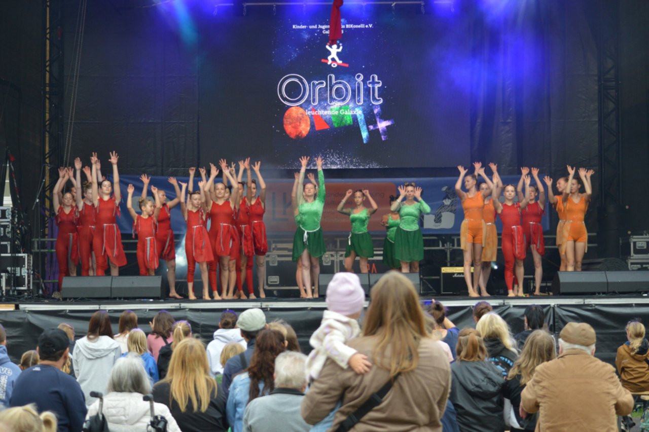 Auf dem Foto ist die Kinder- und Jugendgruppe von Zirkus Bikonelli abgebildet. Die ca. 20 Mädchen präsentieren beim Tag der Niedersachsen auf der LKJ-Bühne ihre Akrobatikkünste. Sie tragen rote und grüne Kostüme und strecken auf dem Bild gerade alle ihre Arme in die Luft. Vor der Bühne stehen und sitzen viele Menschen unterschiedlichen Alters und schauen sich die Show an. 