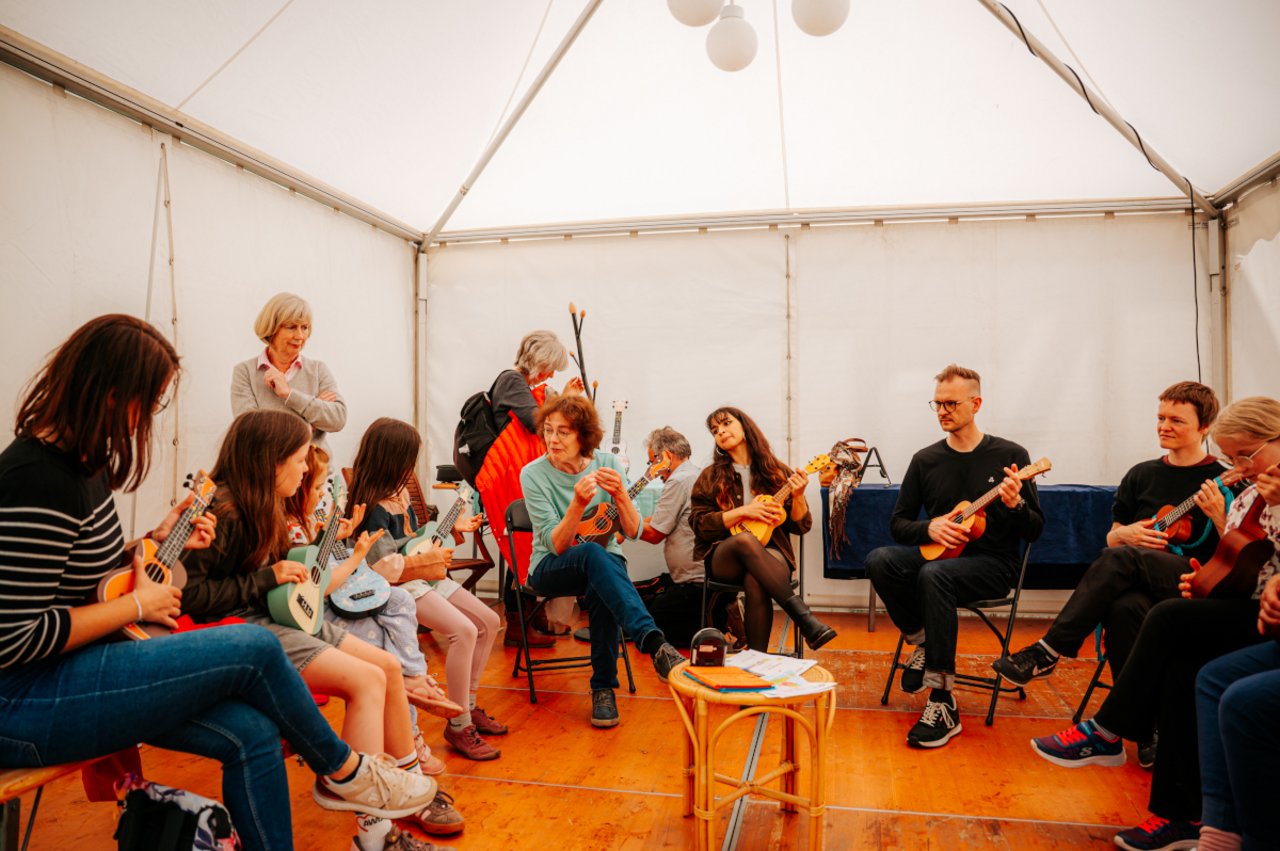 Das Bild zeigt das Innere einer Pagode, in dem Menschen im Halbkreis sitzen. Sie halten Ukulelen in den Händen, während die Workshop-Leiterin Mechthild Werner, die mit im Halbkreis sitzt, etwas erklärt. Beim ›Immer Gute Laune mit Ukulele‹-Workshop haben die großen und kleinen Teilnehmer*innen das Instrument kennengelernt. Das LKJ-Mitglied Deutscher Tonkünstlerverband hat den Kurs organisiert. 